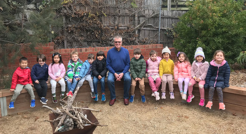 a group of children and man sitting in a row on a bench. The man is in the middle. There is a firepit in front full of sticks and wood.