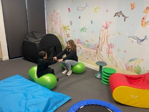 Teacher and student sit opposite each other, with arms outstreched. There are sensory toys and play mats all around them. 