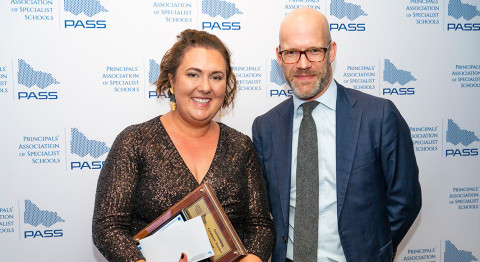 Two people smile at the camera, standing in front of a media wall at the PASS Awards